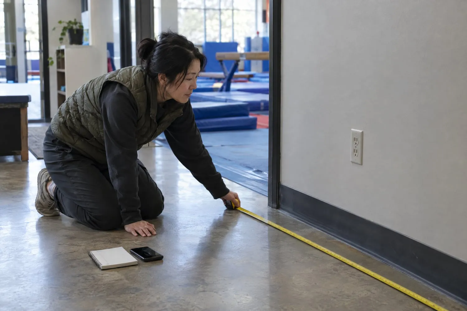 A woman kneels with a tape measure beside a wall outlet in a gym lobby, checking floor space for a vending machine