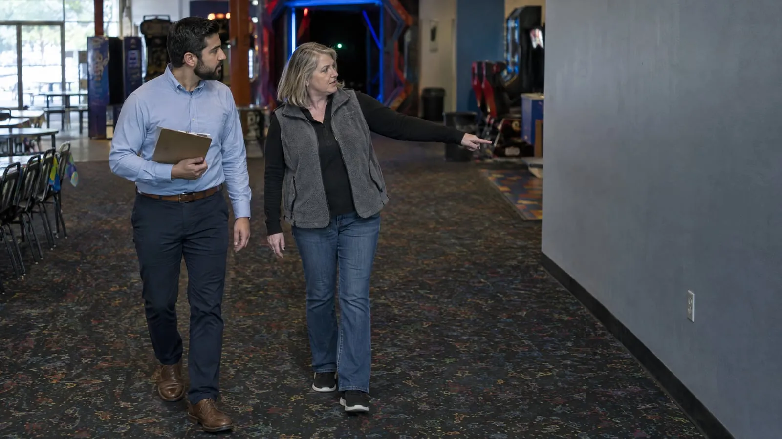 A locator's scout with a clipboard walks a family entertainment center with its manager, who points to an empty wall