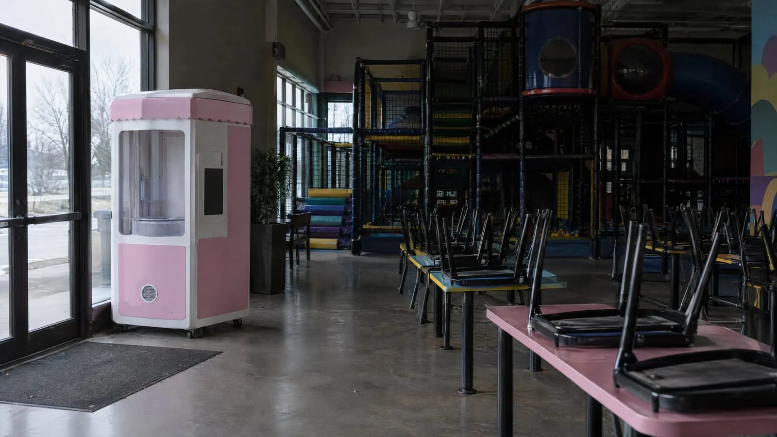 A powered-off cotton candy vending kiosk stands near the entrance of a closed indoor playground, chairs stacked on tables